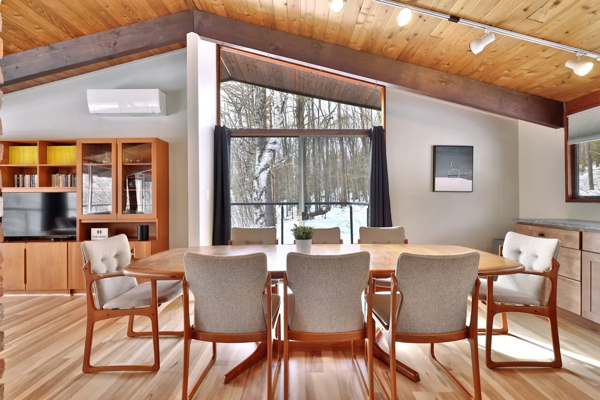 A dining area with a large table, expansive windows letting in natural light, a wooden ceiling, and bookshelves filled with books. The scene is cozy, intimate, and suited for group meals, games, or relaxed conversations while enjoying the view.