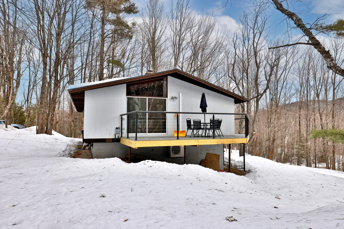 The modern cabin structure is shown with an outdoor seating area on a snow-covered deck, surrounded by trees. The scene suggests relaxation and the opportunity to unwind after a day of adventure, perhaps savoring hot cocoa while watching the snowfall or stargazing at night.