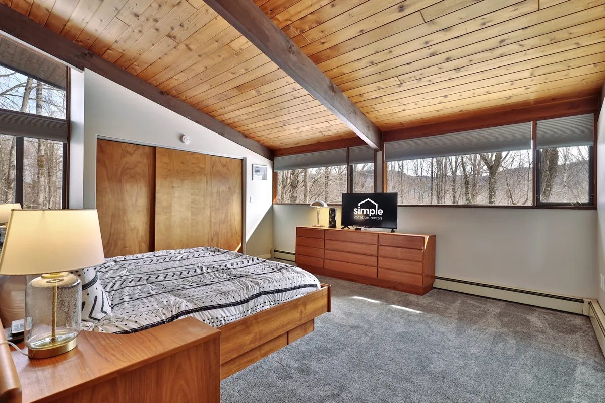 A serene primary bedroom with a king bed adorned in patterned bedding is bathed in natural light from large windows. The room features a modern dresser, a television, and a wooden ceiling that creates a warm, retreat-like atmosphere.