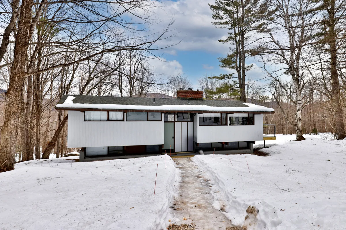 This image showcases the modern architectural design of The Killington Bungalow set amidst a snow-covered landscape and surrounded by trees. The snowy pathway leads directly to the entrance, highlighting the property's connection to the outdoors and the natural beauty of the area throughout the seasons.
