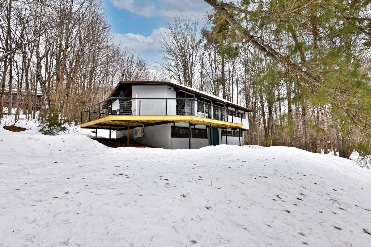 An exterior view featuring a modern house with an elevated deck, surrounded by snow-covered ground and trees. The deck is ideally suited for outdoor grilling and summer BBQs, seamlessly connecting the interior to Vermont’s beautiful outdoors.