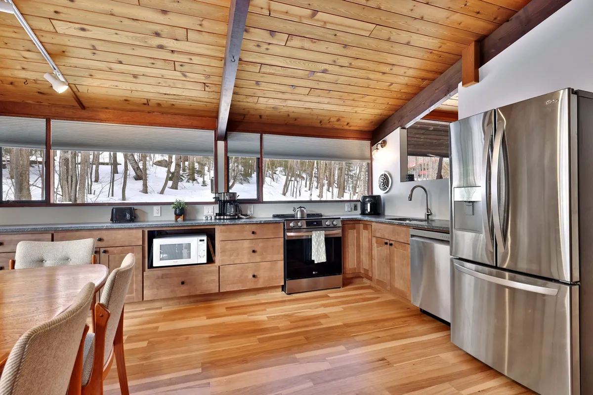 A stylish kitchen-dining space featuring a spacious wooden dining table, large windows with snowy views, a striking wooden ceiling, and sleek stainless steel appliances. The kitchen and dining area are seamlessly connected, with ample natural light enhancing the welcoming atmosphere.