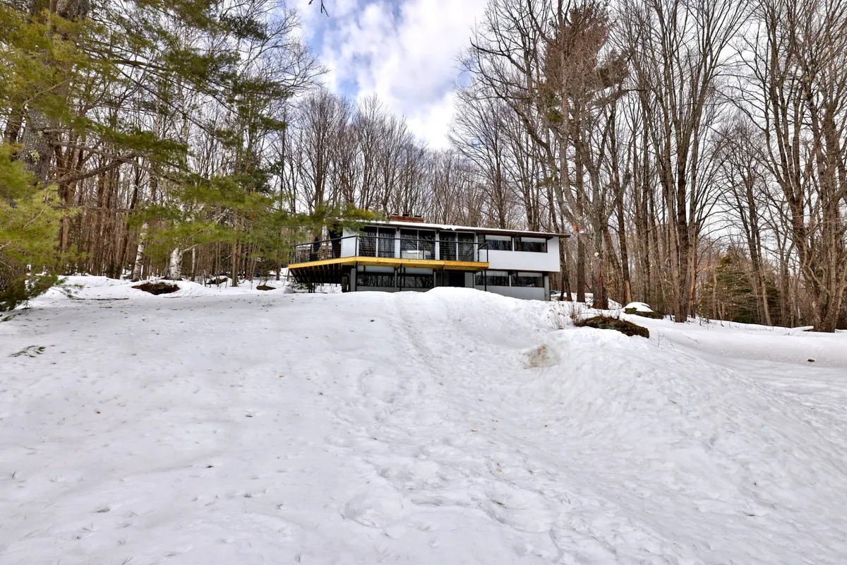 A tranquil exterior view of the house with modern architecture, blanketed by fresh snow and framed by tall trees. The outdoor scene invites guests to imagine themselves stepping out for winter hikes, snowball fights, or simply enjoying the snowy woods nearby.