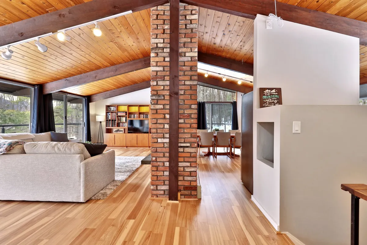 A living area showcasing a warm, welcoming sofa, a brick column, large windows that bring in natural light, a wooden ceiling, and an entertainment unit. The arrangement invites guests to unwind, stream their favorite shows, or enjoy scenic views.