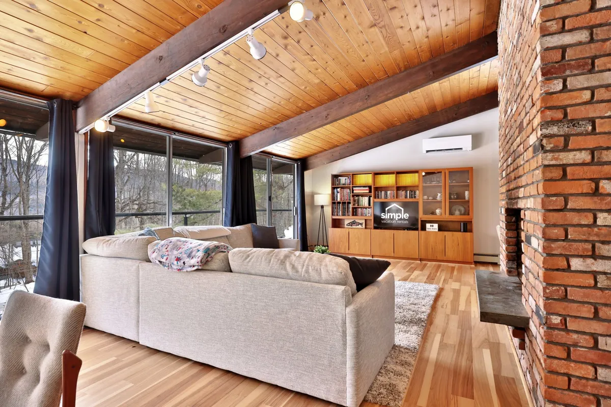 A bright, modern living room featuring a comfortable sectional sofa facing a brick fireplace, with exposed wooden beams overhead and large windows that let in an abundance of natural light. The wooden shelving unit adds warmth and personality to the space, while peaceful wooded views are visible outside the windows.
