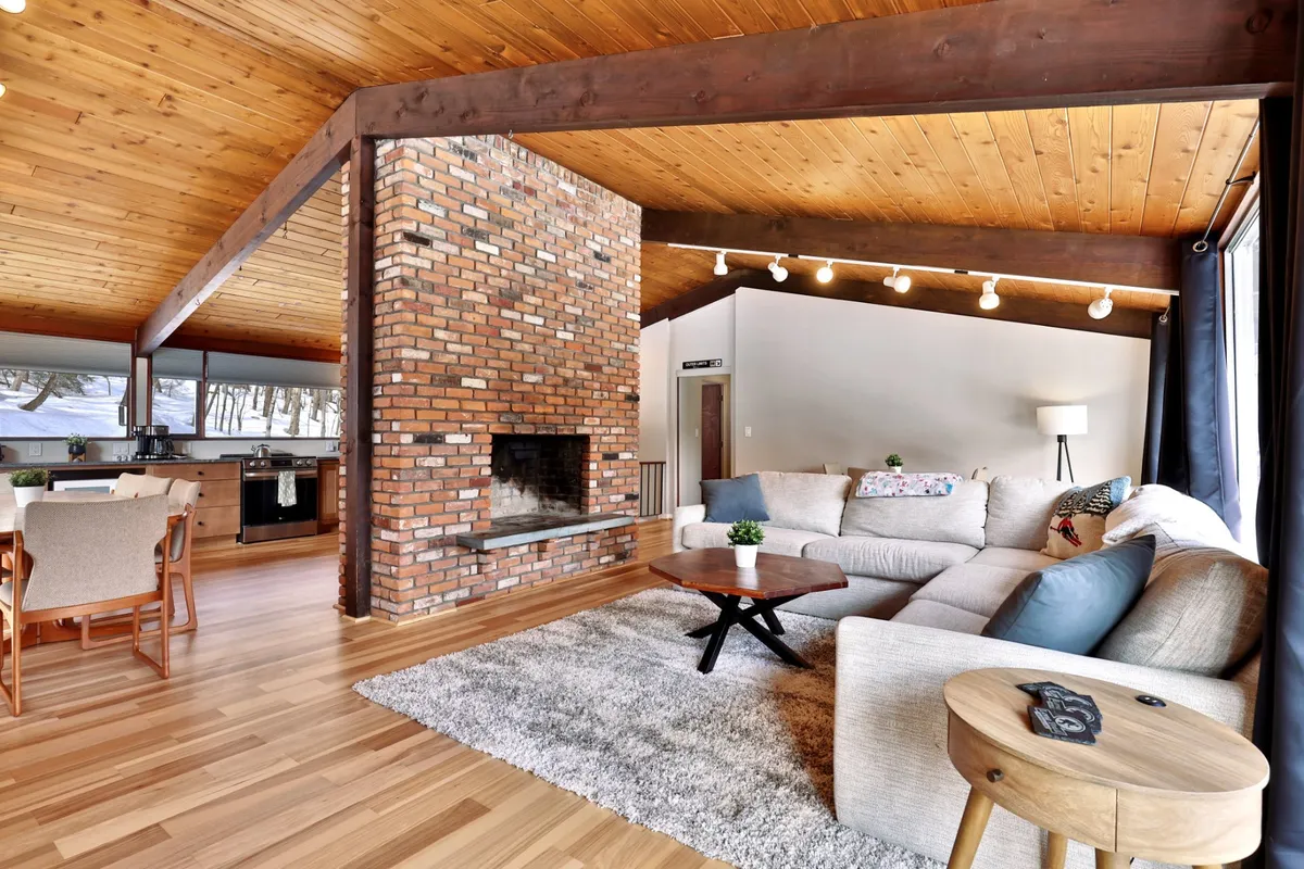 A warm, welcoming interior with a brick fireplace, comfortable sofa, wooden ceiling beams, and an adjacent dining area. The space suggests communal gatherings, relaxation, and shared meals after outdoor activities.