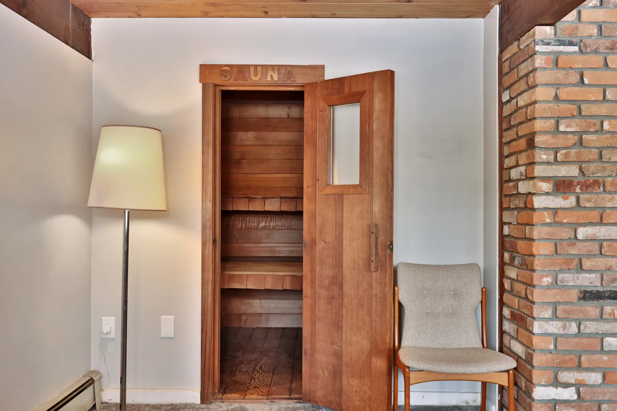 A cozy sauna scene featuring a softly-lit lamp, wood-clad walls, a comfortable chair, and a relaxed, calming atmosphere. The image suggests a space not just for physical wellness but also for mental relaxation and social moments.