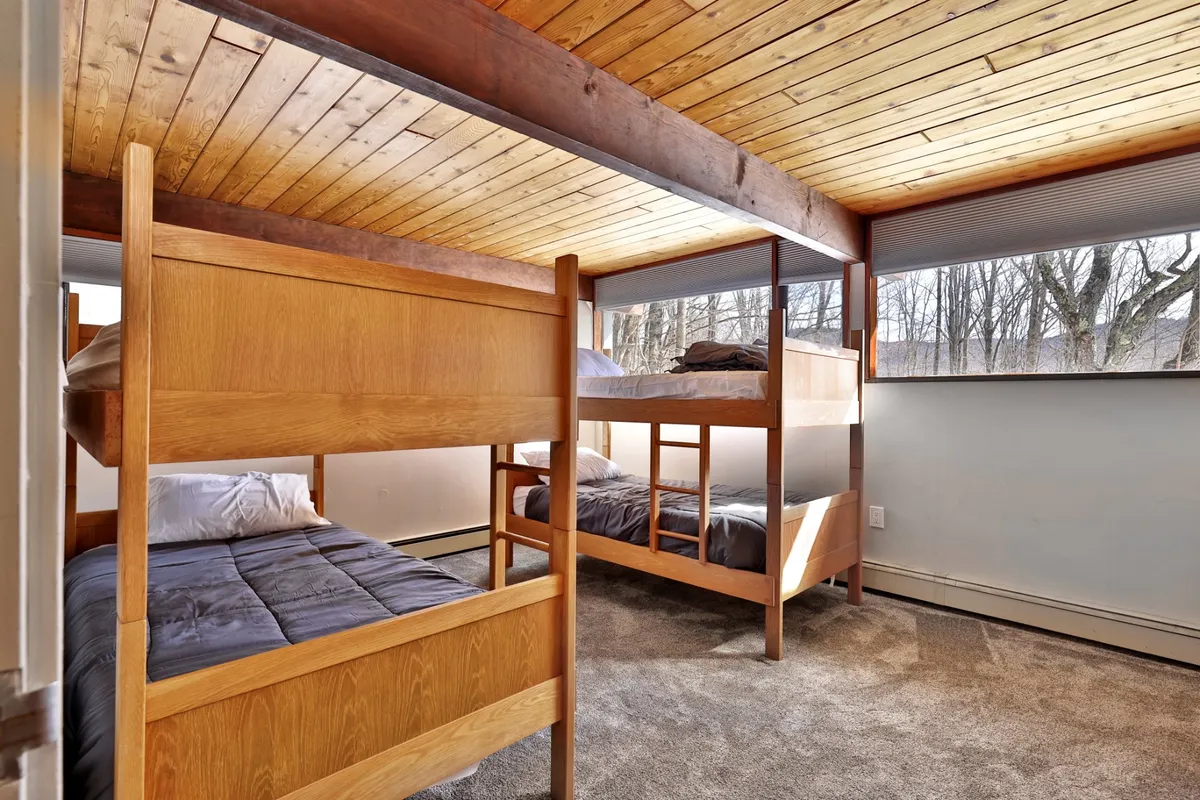 A cozy bedroom with two sturdy bunk beds, large windows letting in sunlight, and a wooden ceiling overhead. The layout is inviting and practical, ideal for families or groups with children.