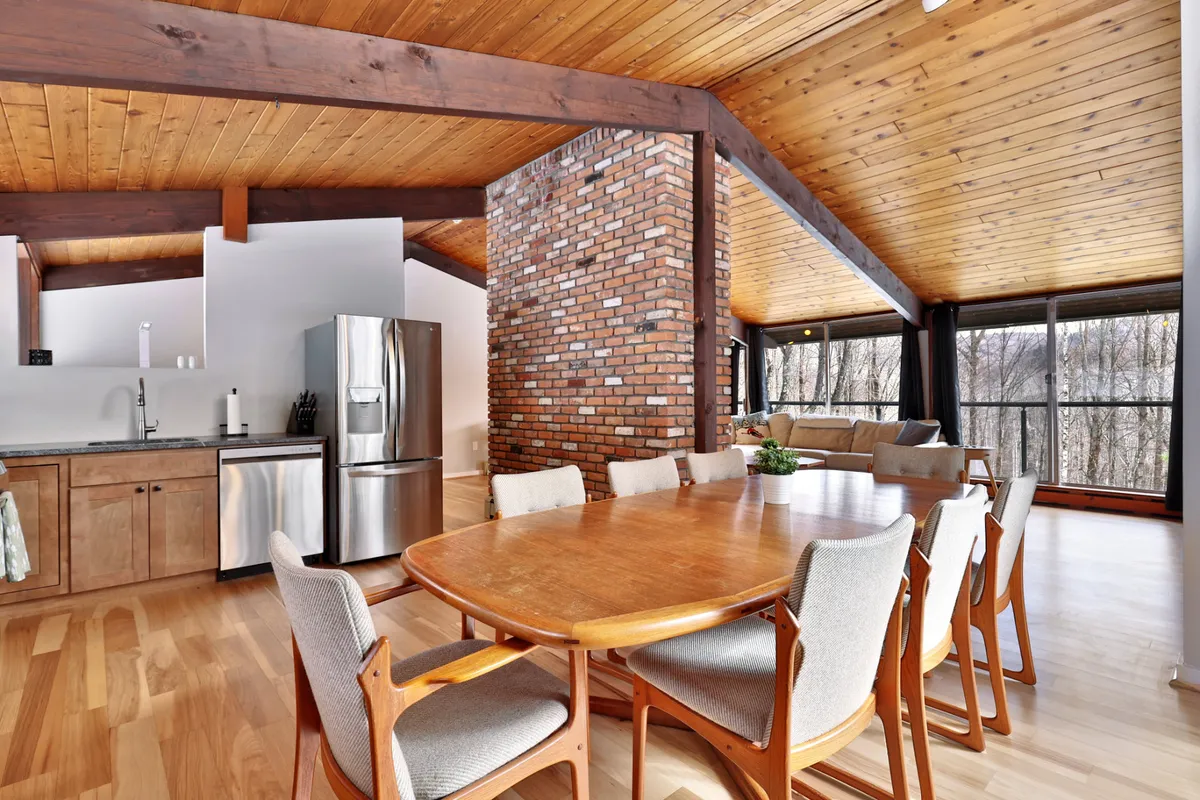 A spacious dining table set against a brick wall and flanked by large windows, with the kitchen visible in the background. The scene is staged for a meal or social gathering, emphasizing the communal aspect of dining.