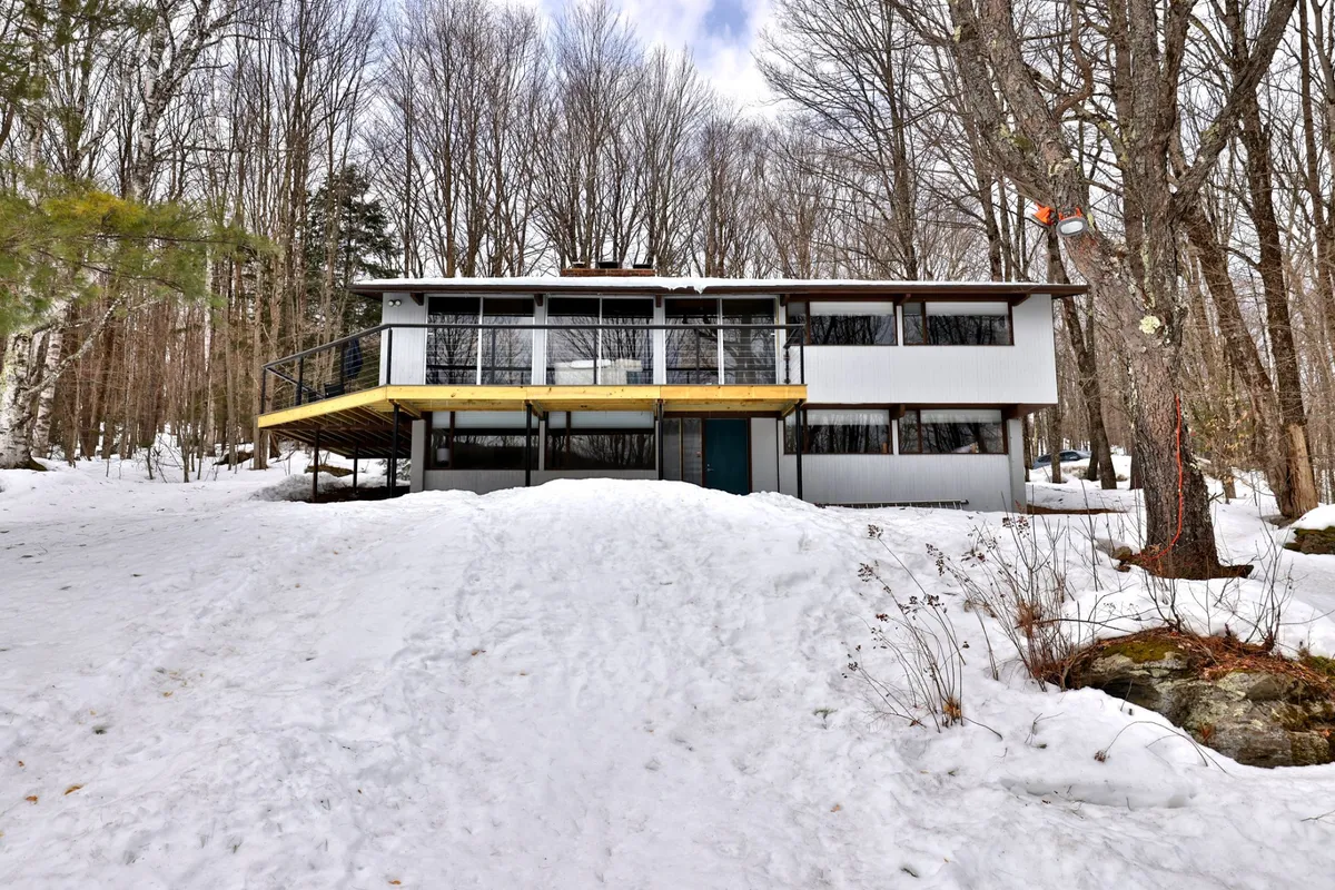An inviting exterior view of the Killington Bungalow nestled among trees, featuring the home’s deck and snow-dusted yard. The scene evokes the potential for outdoor adventures and cozy evenings, with the house set against a tranquil winter backdrop.